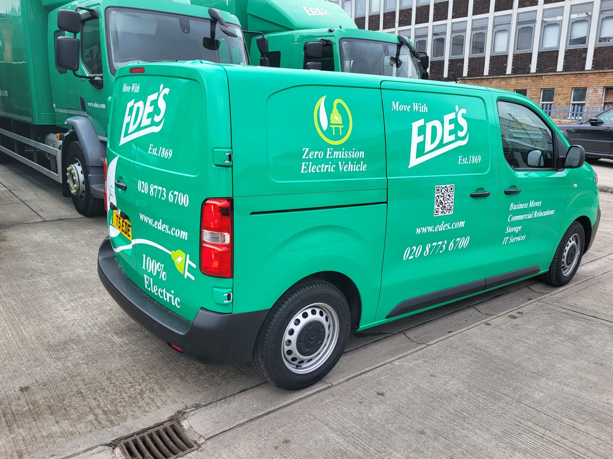 A green van is parked in front of a truck, highlighting its environmentally friendly nature.