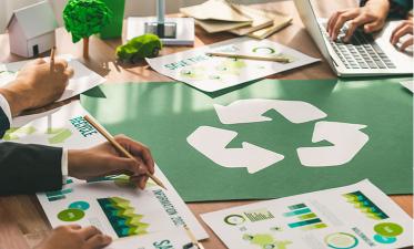 People are working at a table with papers displaying graphs and charts related to sustainability, alongside a laptop and a large recycling symbol.