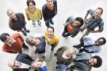 A diverse group of people stand in a circle looking up at the camera, each dressed in casual attire, with various expressions on their faces.