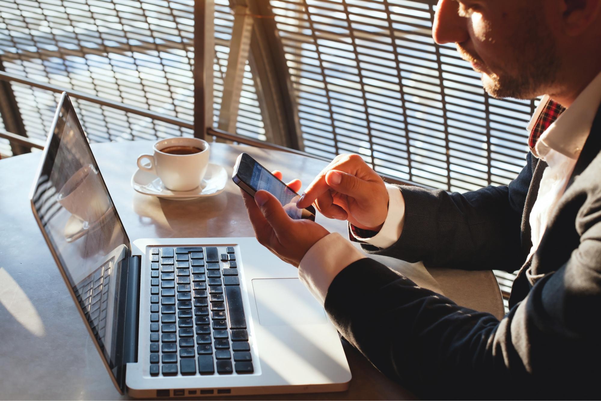 A person in a suit uses a smartphone while sitting at a table with an open laptop and a cup of coffee.