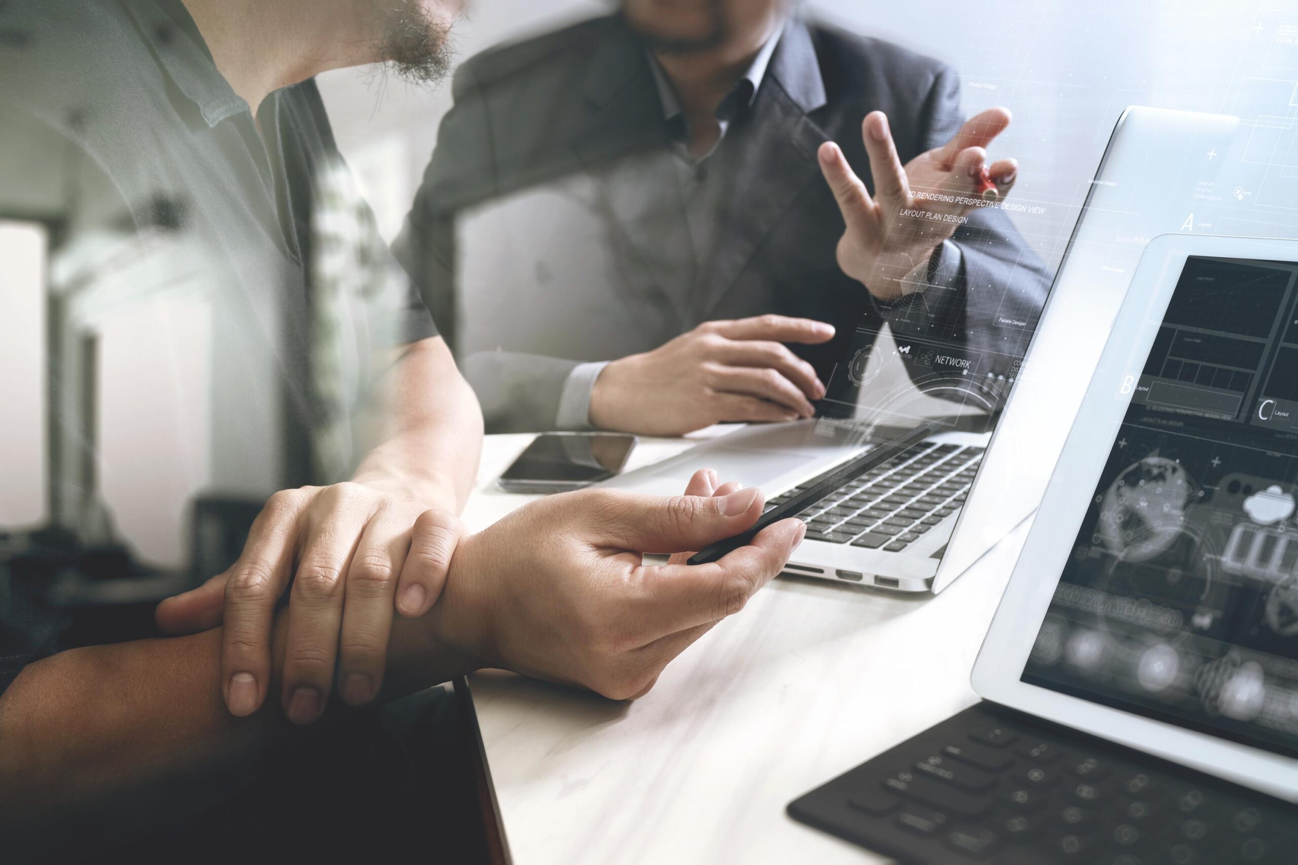 Two people in a meeting, discussing something over a laptop and a tablet with documents and visuals on the screens. One person gestures with their hand while the other holds a pen.