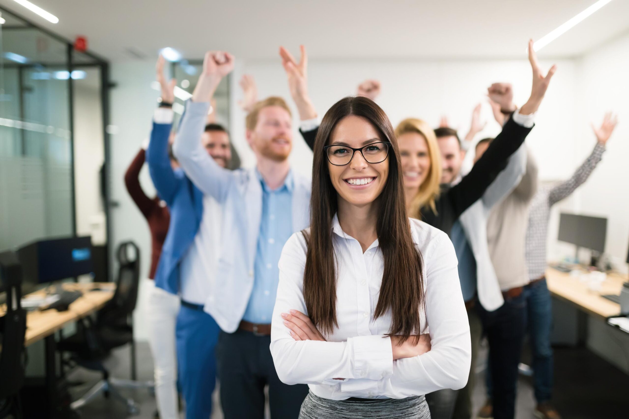 A woman with glasses stands smiling with arms crossed in front of a group of cheering colleagues in an office setting.